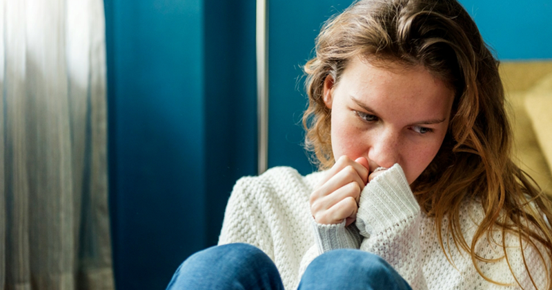 A teenage girl sits on a couch with her hands to her mouth, looking worried