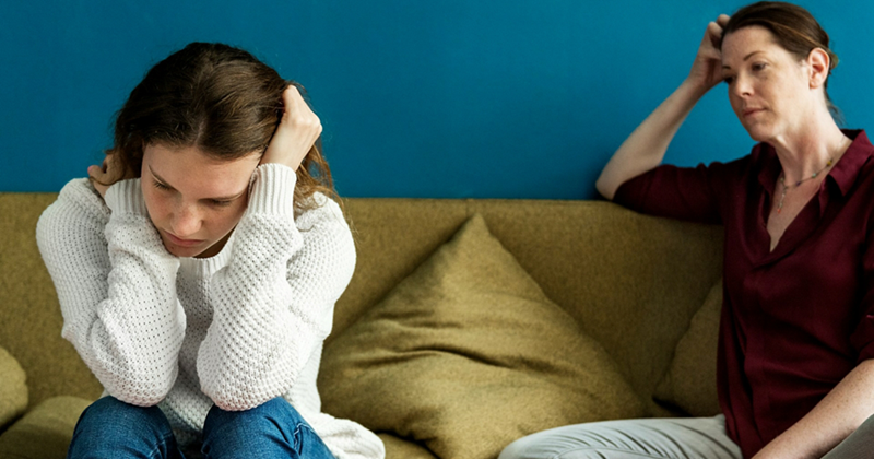 A teenage girl sits on a couch with her head in her hands, an older woman looking displeased beside her