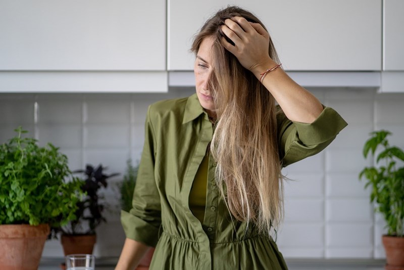 Woman in a green shirt standing in a kitchen with one hand in her hair, surrounded by potted herbs.