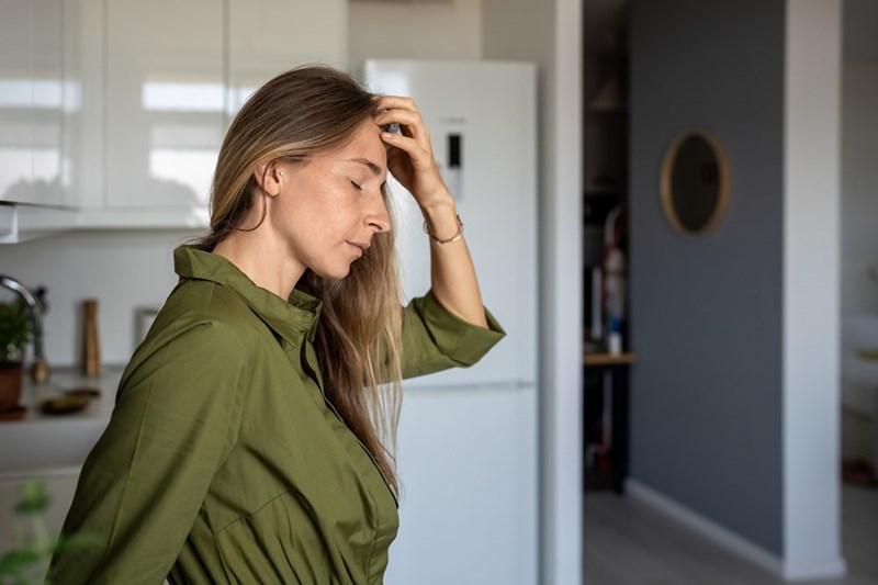 Woman in a green shirt stands in a kitchen with eyes closed and one hand on her head.