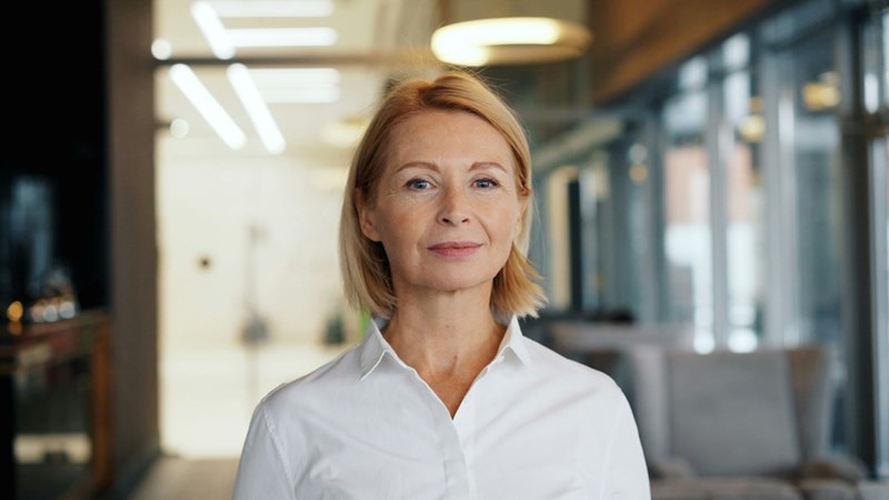 An older businesswoman, dressed in business professional attire, looks out with a smirk, ready to talk.