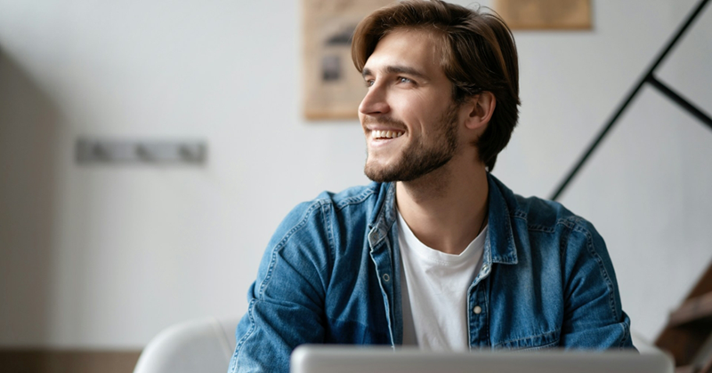 A man sitting with a laptop looks to the side and smiles