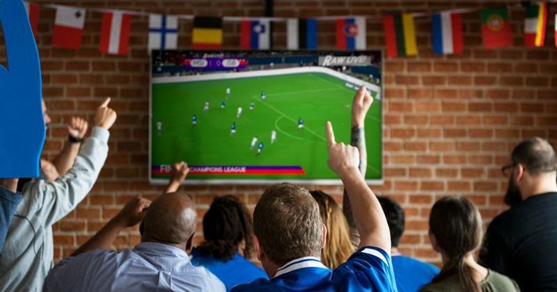 A group of fans cheers as they watch a soccer match on a flat screen TV in a bar
