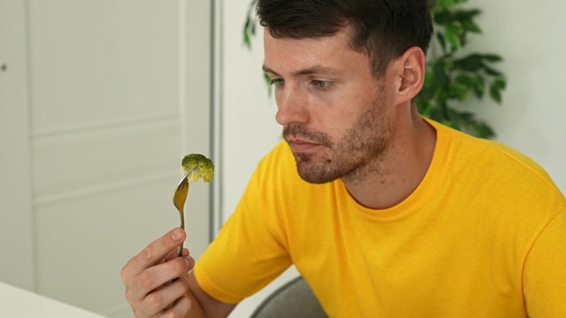 Man in a yellow shirt holding a fork with broccoli, looking down at the food indoors.