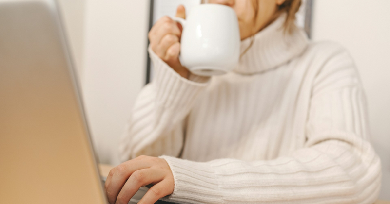 A close-up of a woman drinking from a mug while working on her laptop