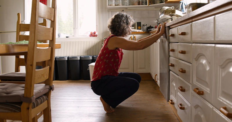 An older woman holds on to the kitchen counter as she crouches in front of the dishwasher