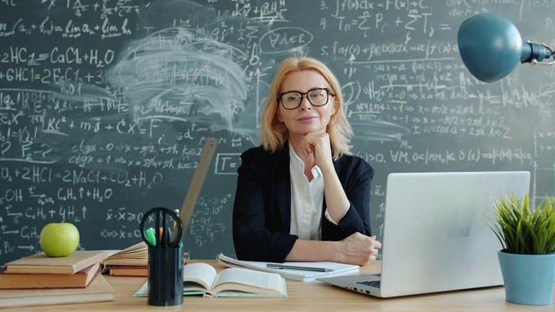 woman in glasses at a desk smiling with a chalkboard full of equations behind