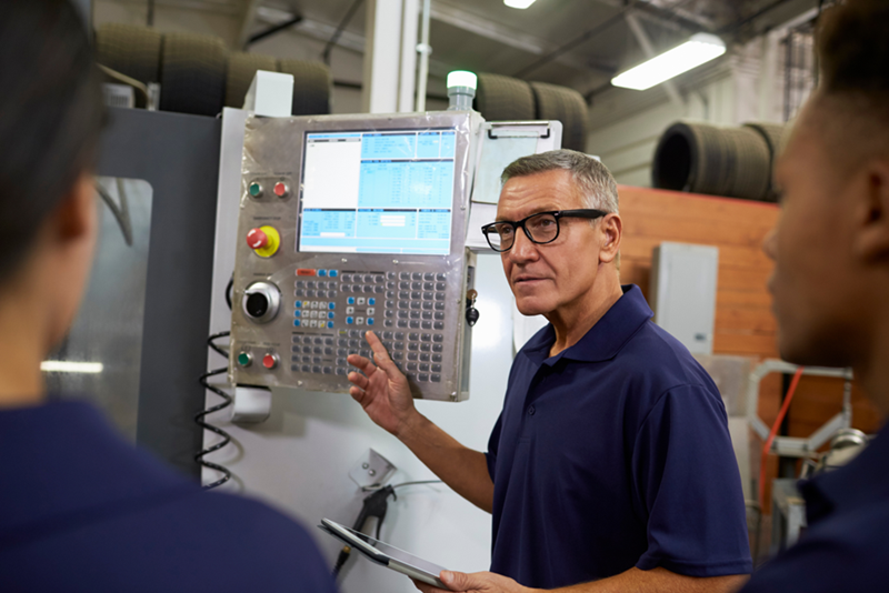 Man holding a tablet beside an industrial control panel while speaking to two people in a workshop.
