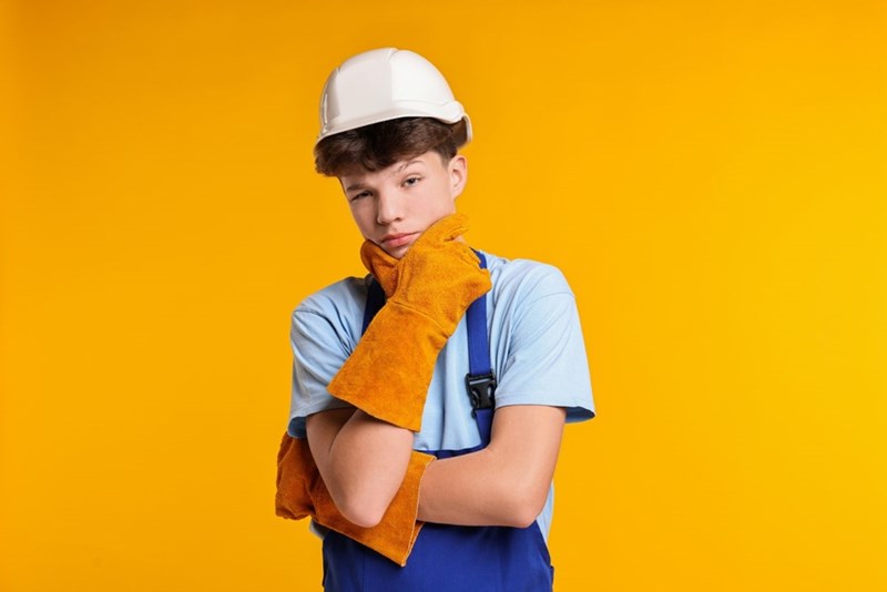Young worker in a hard hat and orange gloves poses against a bright yellow background.