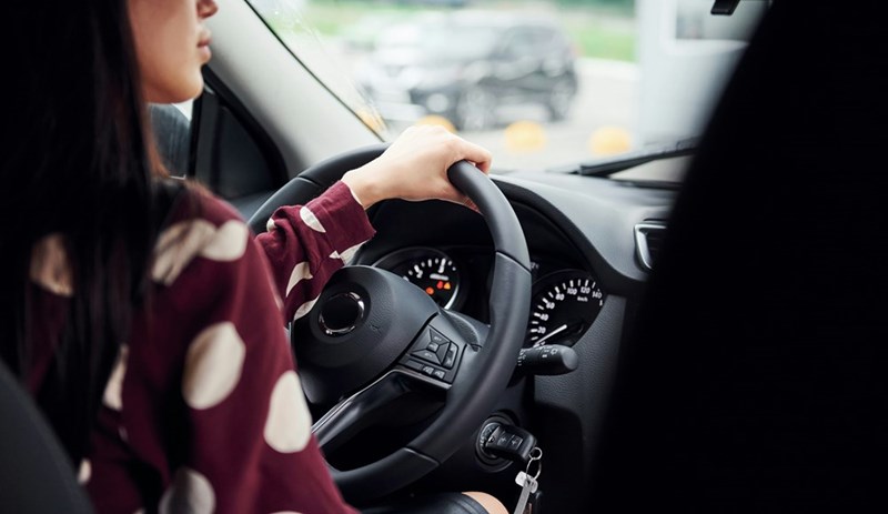 Woman driving a car with one hand on the steering wheel, viewed from the passenger side.