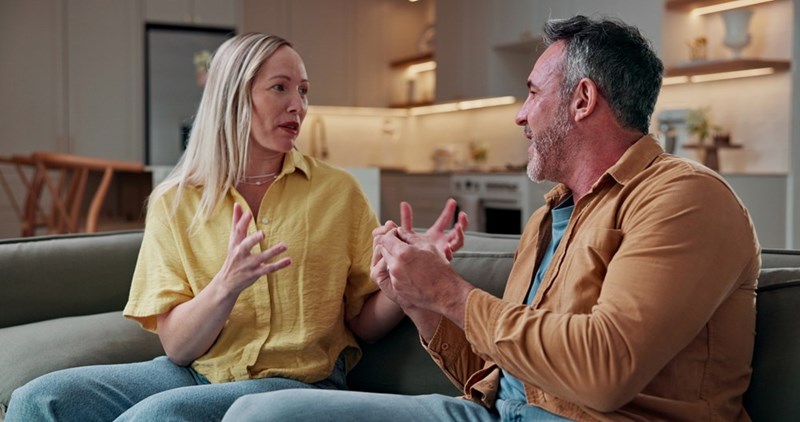 Woman in a yellow shirt gestures while talking with a man on a couch in a living room.