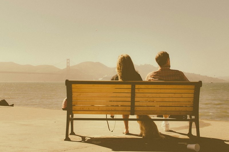 A man and a woman sitting on a bench beside a body of water, model image.