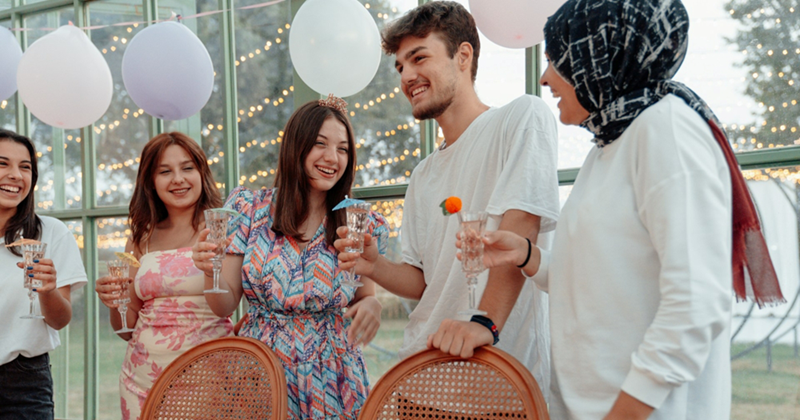 A representation of four women and men standing together holding drinks under a garland of balloons