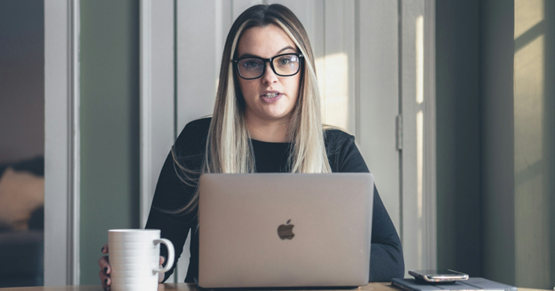 A representation of a female manager sitting behind a MacBook while wearing glasses and holding a mug beside her