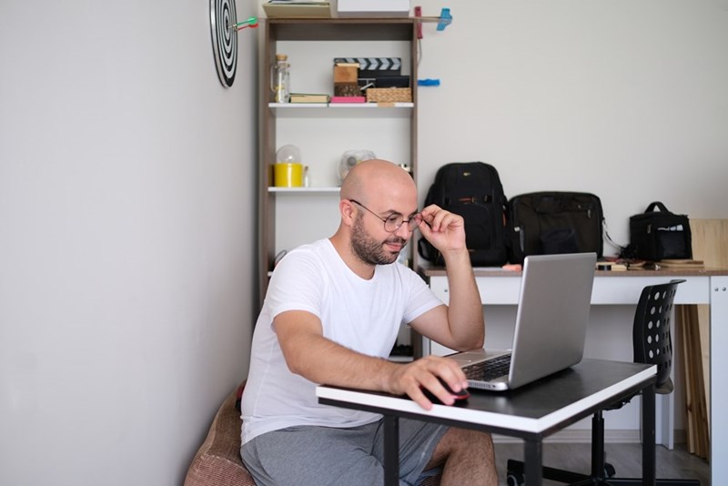 Man in a white T-shirt and gray shorts working on a laptop at a small desk in a home office.
