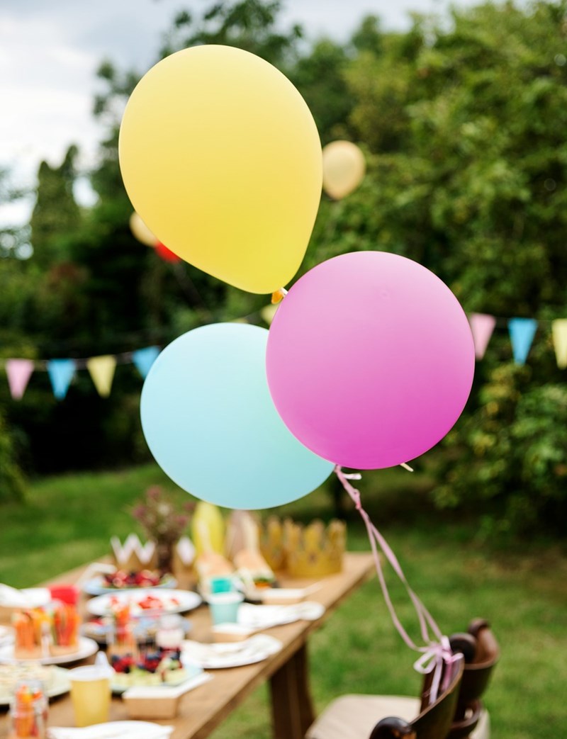 Colorful balloons tied to a chair flow in the wind in the background of an outdoor birthday party.