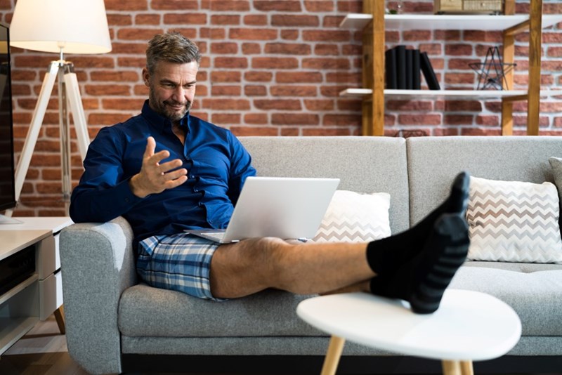 Man sitting on a couch with a laptop, wearing a blue shirt and plaid shorts with feet raised.