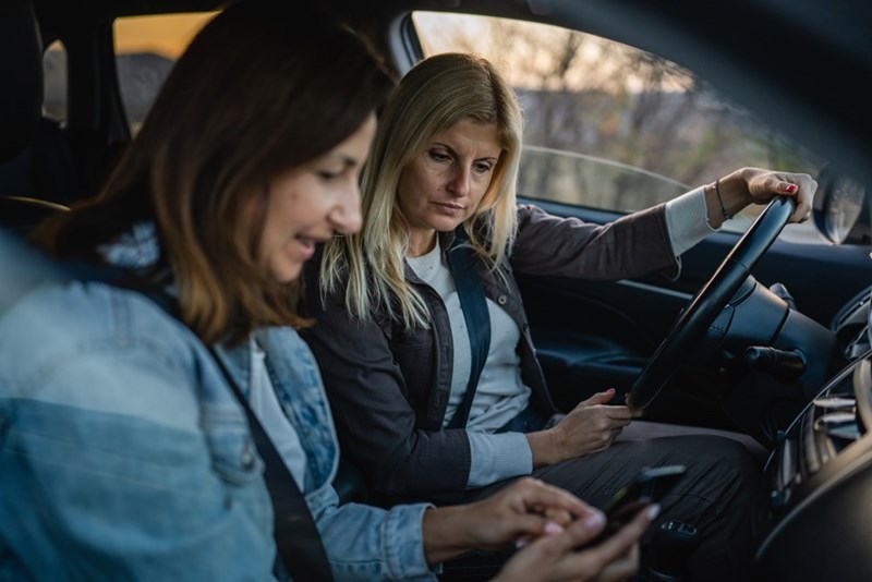 Two women sitting in a car, with the driver looking serious and the passenger holding a phone.