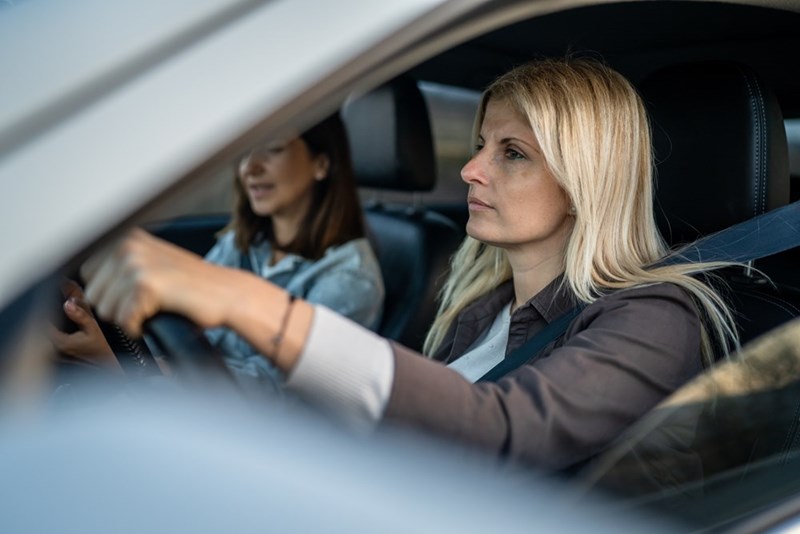 Blonde woman driving a car with a seatbelt on while a passenger sits blurred in the background.