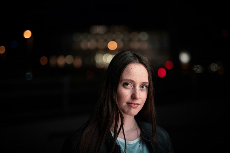 Young woman standing outdoors at night with blurred lights behind her, wearing a dark jacket.