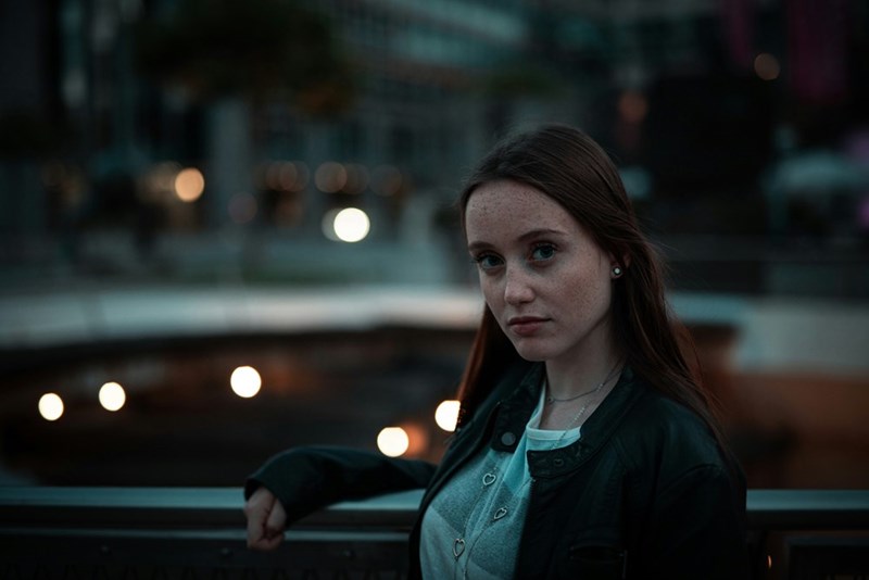 Young woman leaning on an outdoor railing at night, wearing a dark jacket with blurred city lights behind her.