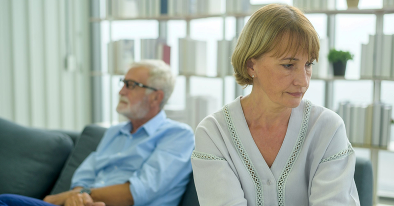 A representation of an older woman looking away with an older man sitting on the couch behind her in the background