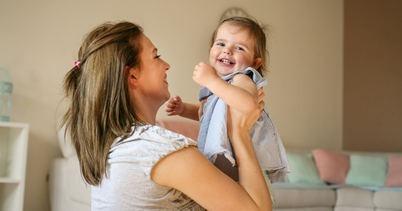 A toddler smiles as a woman lifts her up in her room