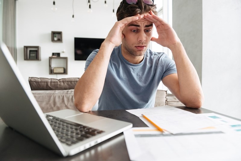Man at a table with a laptop and papers, holding both hands to his forehead with a worried expression.