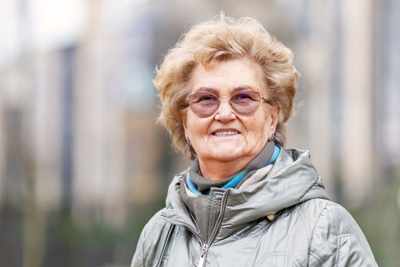Older woman wearing tinted glasses and a silver jacket outdoors with a blurred background.