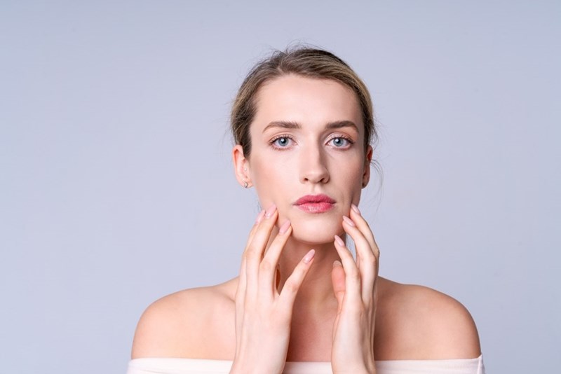 Young woman with hands near her face, wearing an off-shoulder top against a light gray background.