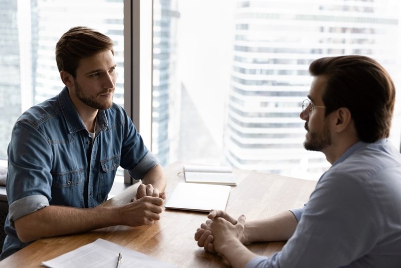 Two men sitting at a conference table in a bright office, having a serious conversation near large windows.
