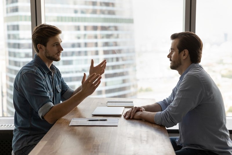 Two men sit at a conference table in a bright office, one gesturing during a serious conversation.