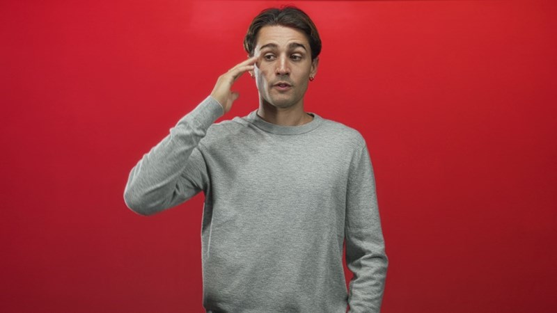Man in a gray sweatshirt raises a hand to his temple while standing against a red background.