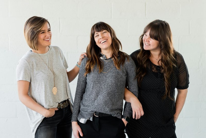 Three women laughing together against a plain wall, like a partnership before trust starts to crack, as shown by models.