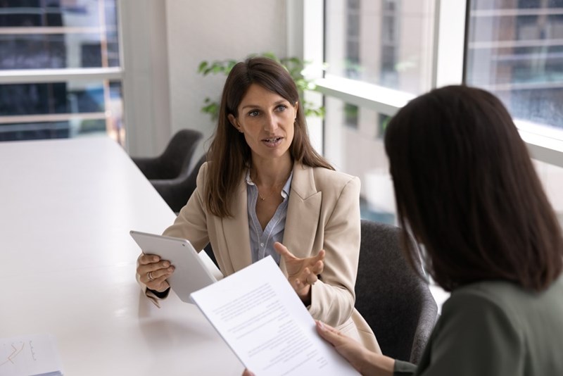 Woman in a blazer speaking across an office table with a controlled, frustrated look