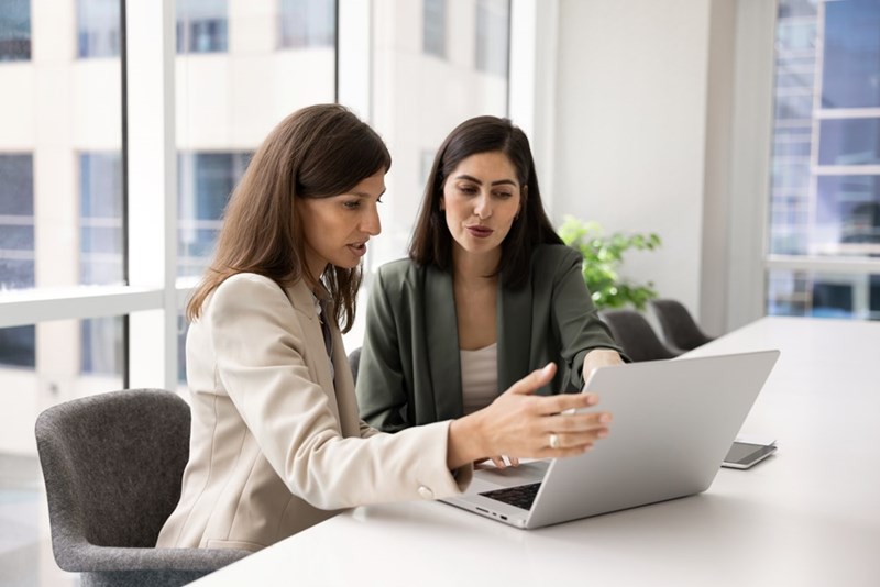 Two women in blazers sit at a laptop in a bright office, discussing something on the screen.