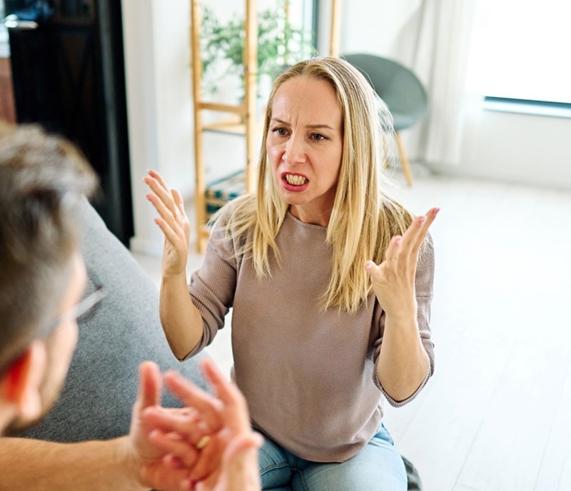Woman gestures angrily while speaking to a man on a couch in a bright living room.
