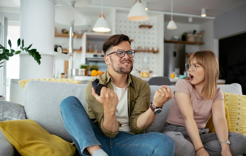 Man holding a game controller on a couch while the woman beside him reacts in surprise or disappointment, as shown by models.