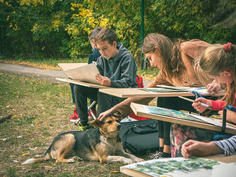 Children drawing outdoors with a dog nearby, illustrating the playful and unconventional childcare style of a family’s chaotic young nanny.