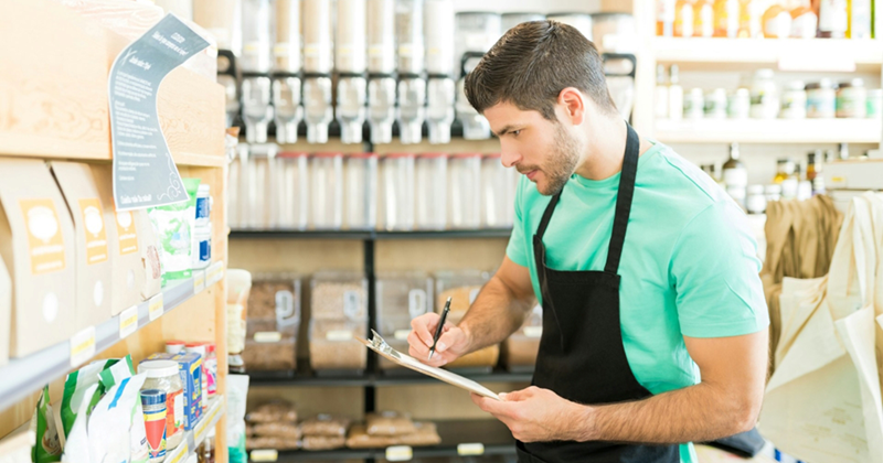 A retail worker writes on a clipboard while looking at shelves of groceries