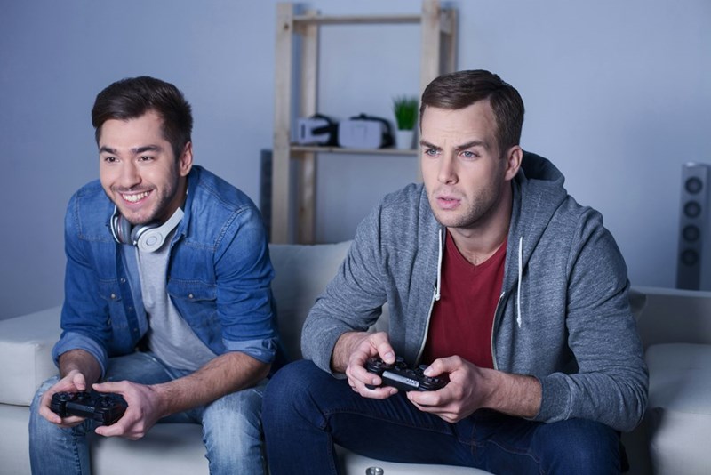 Two gamers playing video games together, sitting on a couch, picturing a relationship tested by dishonesty and unfair play in video games.