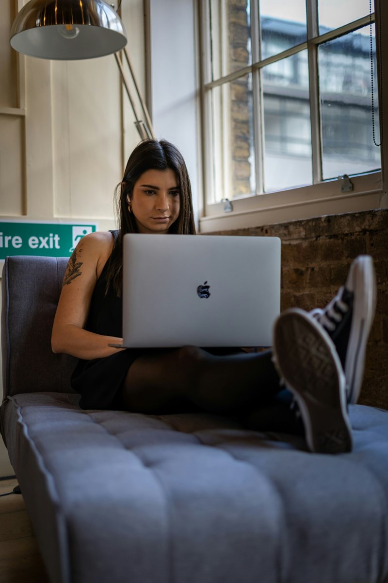 Woman sitting on a gray lounge chair with a laptop near tall windows and a floor lamp.