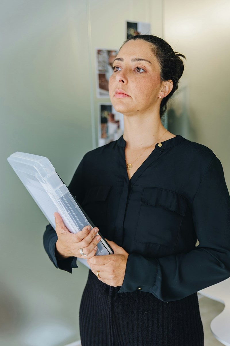 Woman in black holding a clear file case indoors, looking serious beside a pale wall.