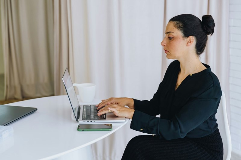 Woman in black typing on a laptop at a white table, with a phone and mug nearby.