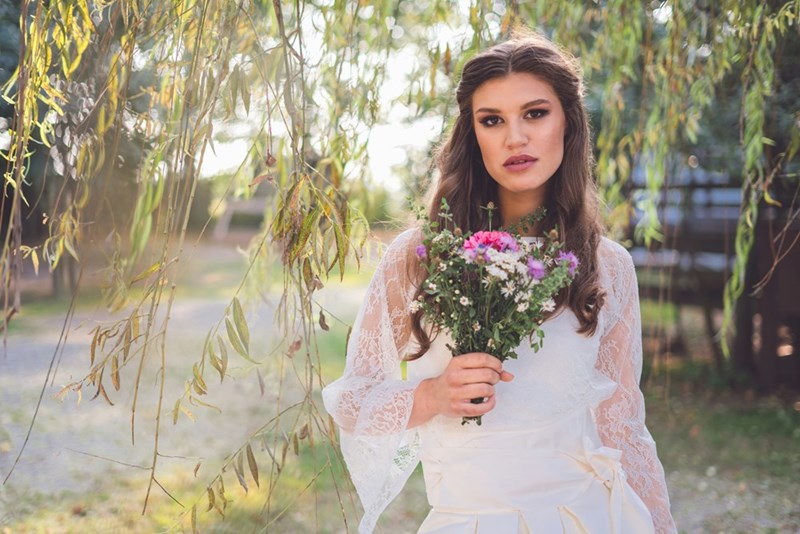 Woman in a white lace dress holding a bouquet outdoors, looking serious beneath hanging tree branches
