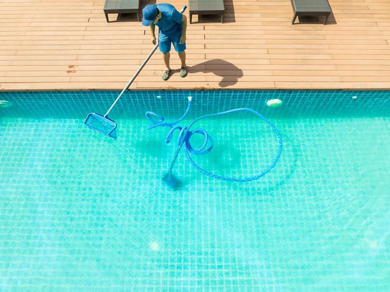 Pool worker uses a long skimmer net to clean a bright blue swimming pool from a wooden deck.