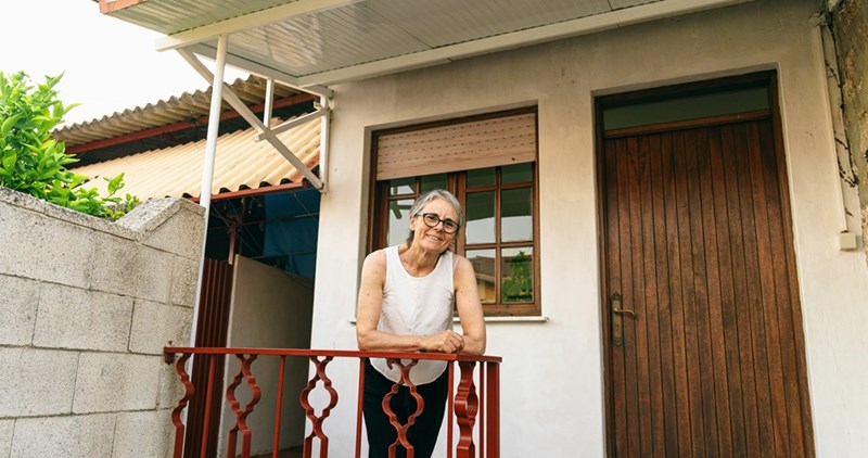 Older woman wearing glasses leans on a red porch railing outside a house with a wooden door.
