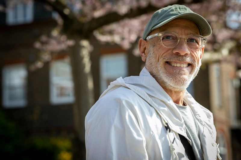 Older man in a cap and glasses smiles outdoors in front of trees and a blurred building.