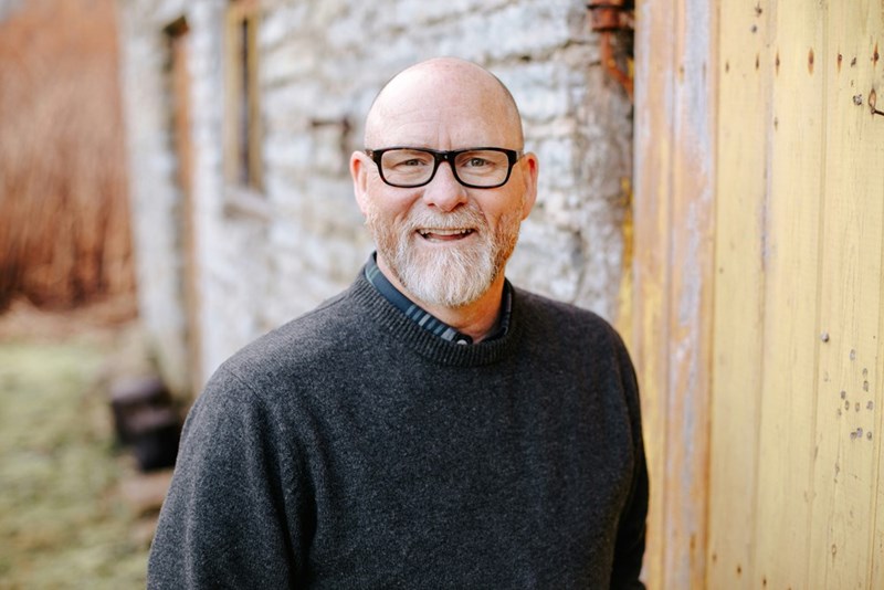 Older man in glasses smiles beside a wooden door and stone wall, wearing a dark sweater.