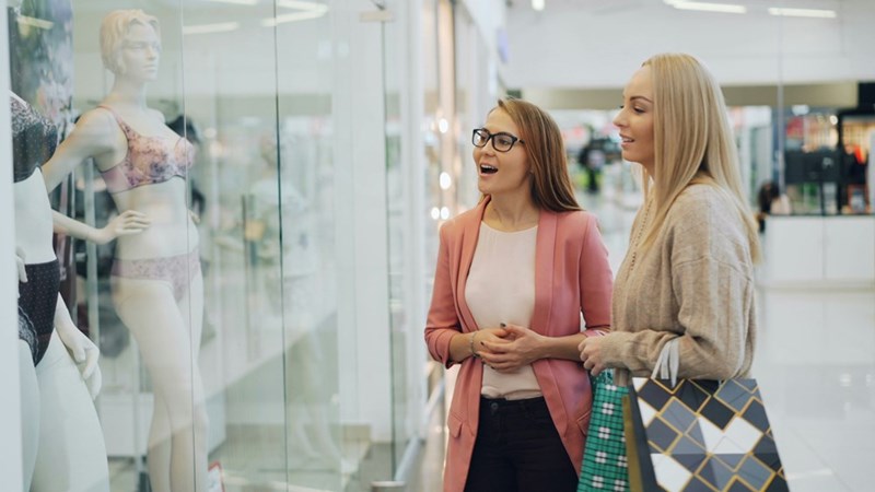 Two retail shoppers in cardigans look at a glass window display in a mall at mannequins